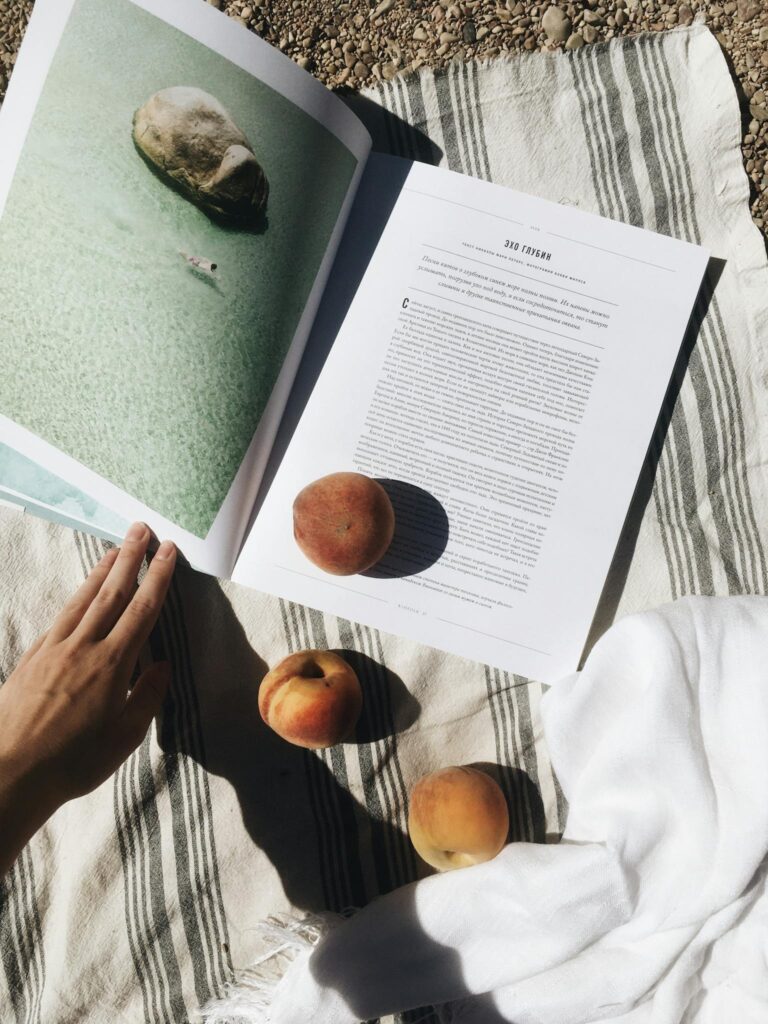 From above of crop unrecognizable traveler reading interesting book while relaxing on beach with ripe peaches on sunny day